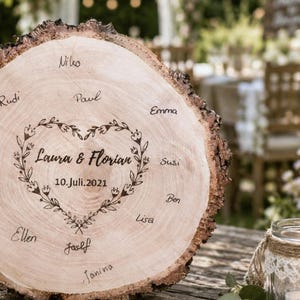 May include: A rustic wooden wedding sign with a natural bark edge. The sign is engraved with the names "Laura & Florian" inside a floral heart, along with the date "10. Juli.2021". Names of guests are written around the edge.