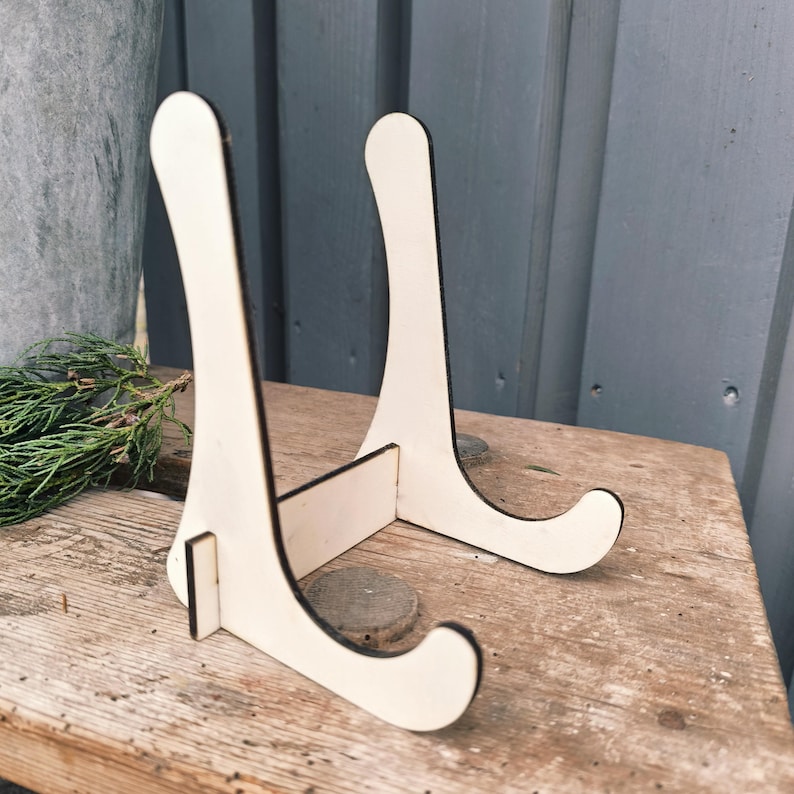 May include: A pair of unfinished wooden display stands. Each stand has a curved top and base, connected by a horizontal support. The stands are made of light-colored wood with dark edges, suitable for displaying plates or artwork.