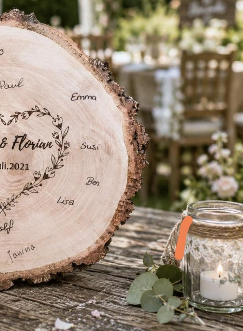 May include: A rustic wooden wedding sign with names and the date "Juli.2021" engraved on it. A small glass jar with a lit candle and twine detail sits next to the sign. The background is blurred, suggesting an outdoor event.