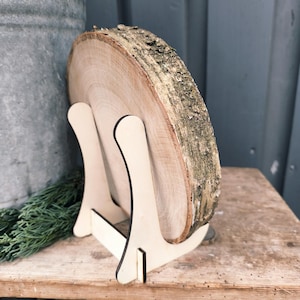 May include: A natural wood slice with bark edges, displayed on a light-colored wooden easel. The wood slice is round and shows the tree's growth rings. The easel has a simple, minimalist design, perfect for displaying art or decor.