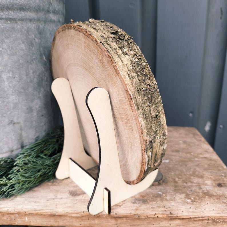 May include: A natural wood slice with bark edges, displayed on a light-colored wooden easel. The wood slice is round and shows the tree's growth rings. The easel has a simple, minimalist design, perfect for displaying art or decor.