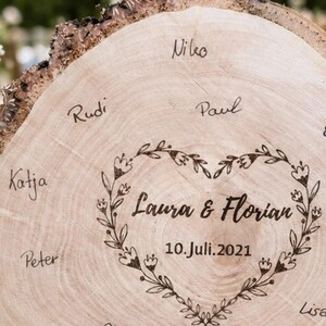 May include: A light brown wooden slice with a natural bark edge. The center features a heart-shaped floral design with the names "Laura & Florian" and the date "10.Juli.2021". Several names are handwritten around the heart.