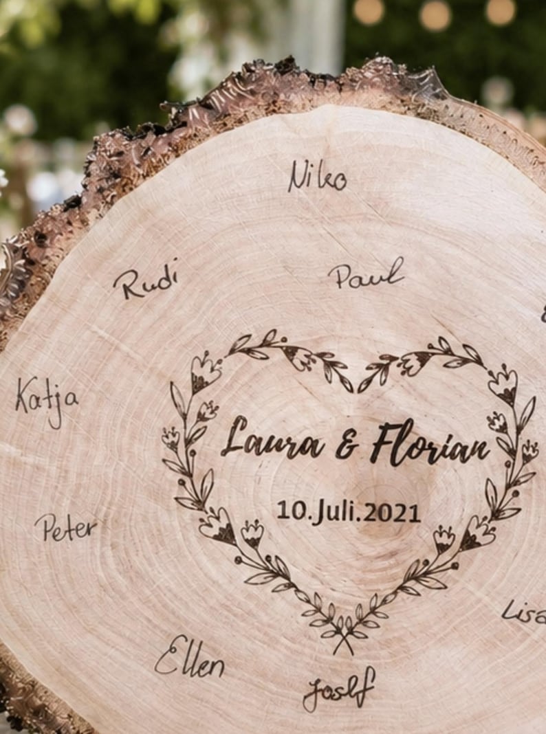 May include: A light brown wooden slice with a natural bark edge. The center features a heart-shaped floral design with the names "Laura & Florian" and the date "10.Juli.2021". Several names are handwritten around the heart.