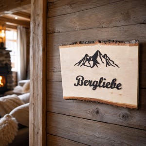Könnte beinhalten: Ein rechteckiges Holzschild mit natürlicher Baumkante, das eine schwarze Silhouette eines Gebirges und das Wort Bergliebe in einer schwarzen Schreibschrift zeigt. Die helle Holzmaserung bildet einen Kontrast zu den dunklen Elementen.
