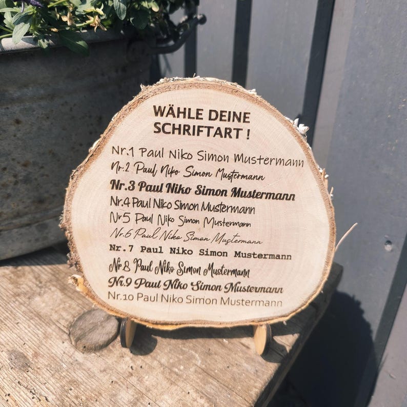 May include: A wooden slice with the text "W&Auml;HLE DEINE SCHRIFTART!" and a list of names. The wood has a natural, light brown color with visible grain. The text is in black, and the wood slice is propped up on small wooden supports.