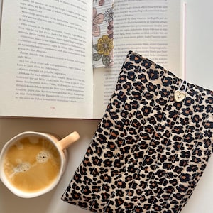 May include: A quilted fabric book sleeve featuring a brown and black leopard print pattern, a white heart-shaped button closure, and a small Nalu brand tag. A matching floral bookmark rests inside an open book next to a mug of coffee.