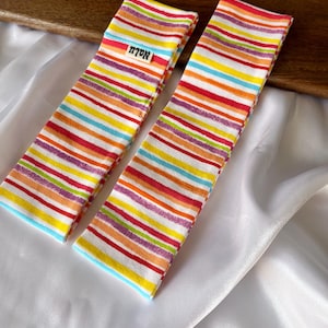 Könnte beinhalten: Zwei Stirnbänder mit bunten Streifen. Die horizontalen Streifen sind in Rot, Orange, Gelb, Blau und Lila gehalten. Auf jedem Stirnband befindet sich ein Etikett mit dem Wort "Nalu". Die Stirnbänder liegen auf einem weißen Satinstoff.