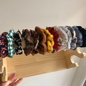 Puede incluir: Un estante de madera con una variedad de scrunchies coloridos, incluyendo rojo, verde, negro, blanco y marrón. Los scrunchies están dispuestos en una fila en el estante.