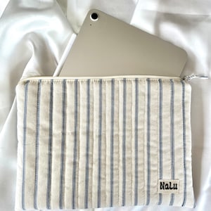 以下が含まれることがあります： クリーム色のタブレットスリーブ。ネイビーブルーのストライプが縦に入っています。スリーブにはジッパー開閉があり、「Nalu」と書かれた小さな長方形のラベルが付いています。シルバーのタブレットとスタイラスがスリーブ内側に部分的に見えます。
