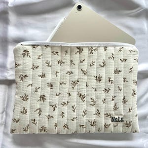 Può includere: Custodia per tablet trapuntata color crema con motivo a foglie marroni. La custodia ha una cerniera bianca e una piccola etichetta con la scritta "Nalu". Un tablet e una penna stilo sono parzialmente visibili.