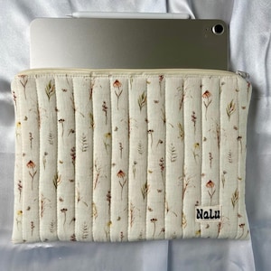 Può includere: Custodia per tablet trapuntata color crema con motivo floreale, contenente un tablet e uno stilo argentati. La custodia ha una chiusura a cerniera e la parola "Nalu" è stampata in basso a destra. Il design floreale include accenti marroni, verdi e gialli.