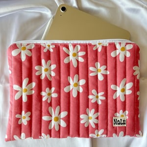 Può includere: Una custodia per tablet rosa trapuntata con stampa di margherite bianche e la parola "Nalu" ricamata nell'angolo in basso a destra.