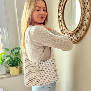 May include: A white tote bag with a black heart pattern and the word "Nalu" printed on it. The bag is carried over the shoulder. The background includes a round woven mirror and a houseplant.