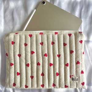 Può includere: Custodia trapuntata color crema per tablet con motivo a cuori rossi. All'interno sono visibili un tablet argentato e una penna. L'etichetta "Nalu" è in basso a destra. Su una superficie di raso bianco.