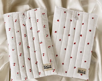 Peut inclure: Deux housses rectangulaires blanches matelassées avec des coutures verticales. L'une présente un motif de cerises rouges, l'autre de fraises rouges. Chacune possède un bouton transparent en haut et une étiquette beige marquée Nalu en bas.
