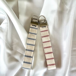 Könnte beinhalten: Zwei Schlüsselanhänger aus Stoff mit Metallbeschlägen. Einer hat blaue horizontale Streifen auf cremefarbenem Grund, der andere rote horizontale Streifen auf cremefarbenem Grund. Beide haben ein kleines Metallschild mit der Gravur Nalu.
