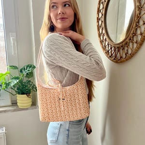 Può includere: Borsa a tracolla color pesca con stampa floreale e cinturino rosa chiaro. La borsa presenta un motivo di piccoli fiori arancioni. La borsa è rettangolare con chiusura a coulisse. La borsa è realizzata in tessuto trapuntato.