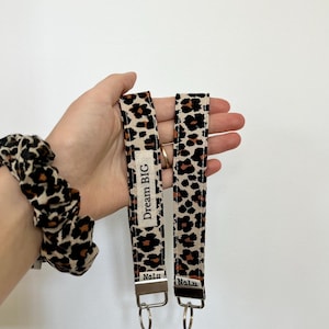 Può includere: Due portachiavi con motivo leopardato, su sfondo bianco. Un portachiavi ha la scritta "Dream BIG" e il marchio "Nalu". Un elastico per capelli abbinato è visibile sullo sfondo.