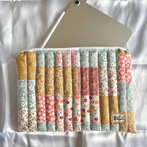 Könnte beinhalten: Eine gesteppte Tablethülle mit Patchwork-Design, das verschiedene Blumenmuster in Gelb-, Grün-, Rot- und Rosatönen zeigt. Die Hülle hat einen Reißverschluss und ein Etikett mit der Aufschrift "Nalu".