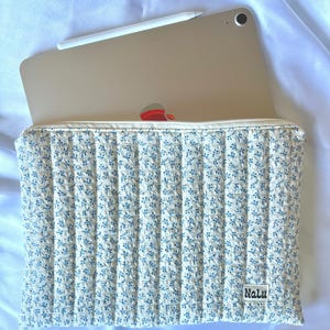 Pode incluir: Uma capa de tablet acolchoada creme com um padrão floral azul. A capa tem um fecho de correr e uma pequena etiqueta que diz "Nalu". Um iPad e uma caneta branca são visíveis no interior.
