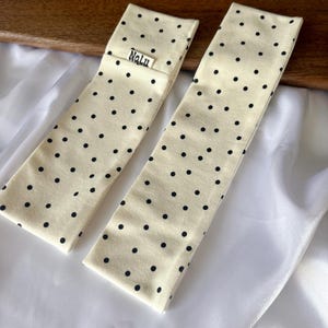 Könnte beinhalten: Zwei cremefarbene Haaraccessoires mit schwarzen Punkten. Eines hat ein kleines Etikett mit der Aufschrift "Nalu". Die rechteckigen Accessoires sind gefaltet und liegen auf einer weißen Satin-Oberfläche.