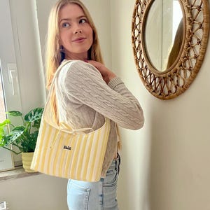 Può includere: Una borsa tote a righe gialle e bianche con la parola "Kalu" ricamata. La borsa ha una chiusura con coulisse color crema e una lunga tracolla. La borsa è portata da una persona.