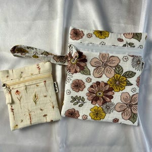 May include: Two small, zippered pouches. One is rectangular with a floral pattern in pink, brown, and yellow on a white background. The other is smaller, rectangular, and has a cream-colored background with a floral pattern. Both have a zipper closure.