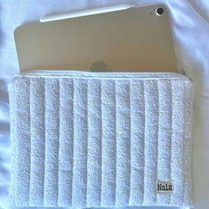 Peut inclure: Une housse blanche matelassée pour iPad avec un motif ajouré floral. L'étui a une fermeture éclair et une petite étiquette avec le mot "Nalu". Un iPad doré et un stylet blanc sont visibles à l'intérieur.
