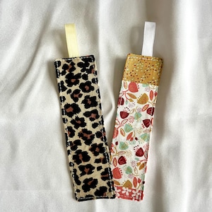 Könnte beinhalten: Zwei Stoff-Lesezeichen. Ein Lesezeichen zeigt ein Leopardenmuster in Braun und Schwarz. Das andere Lesezeichen hat ein Patchwork-Design mit roten, gelben und grünen Blumenmustern.