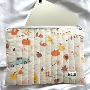 Può includere: Custodia per tablet trapuntata color crema con un motivo a tema autunnale. Il motivo include zucche, foglie, un camion blu e il marchio "Nalu". Un tablet argentato è parzialmente visibile all'interno.