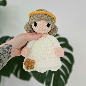 May include: A handmade crochet doll with a white dress, light brown hair, and a yellow headband. The doll has a light pink face and hands, and a brown flower accent on the dress. The doll is approximately 8 inches tall.