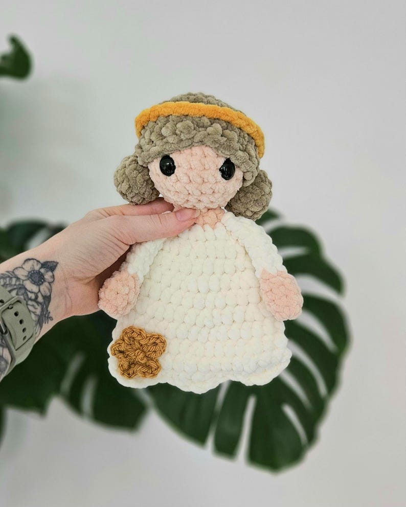 May include: A handmade crochet doll with a white dress, light brown hair, and a yellow headband. The doll has a light pink face and hands, and a brown flower accent on the dress. The doll is approximately 8 inches tall.