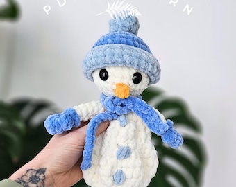 May include: A handmade snowman plushie with a blue hat, scarf, and mittens. The snowman has black eyes and an orange carrot nose. The text "PDF PATTERN" is visible in the background. The plushie is made of soft, textured yarn.