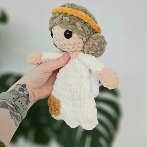 May include: A handmade crochet doll with a halo. The doll has a light skin tone, black eyes, and a white dress. The hair is a light brown color, and the halo is yellow. The doll's hands and feet are a light pink color.