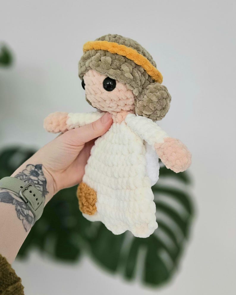 May include: A handmade crochet doll with a halo. The doll has a light skin tone, black eyes, and a white dress. The hair is a light brown color, and the halo is yellow. The doll's hands and feet are a light pink color.