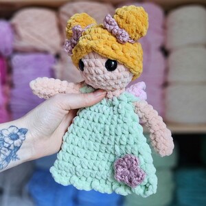 May include: A handmade crochet doll with a light green dress and a yellow hair bun. The doll has black eyes and a pink flower on the dress. The doll is made of soft, plush yarn.