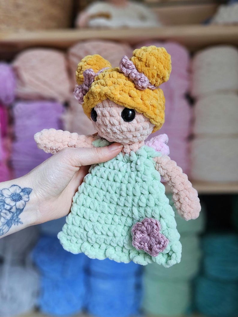 May include: A handmade crochet doll with a light green dress and a yellow hair bun. The doll has black eyes and a pink flower on the dress. The doll is made of soft, plush yarn.