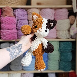 May include: A handmade crochet cat plushie in shades of brown, white, and dark brown. The plush toy has a soft, textured appearance and features a detailed design with small black eyes and a pink nose. The cat is held in front of a shelf filled with yarn.