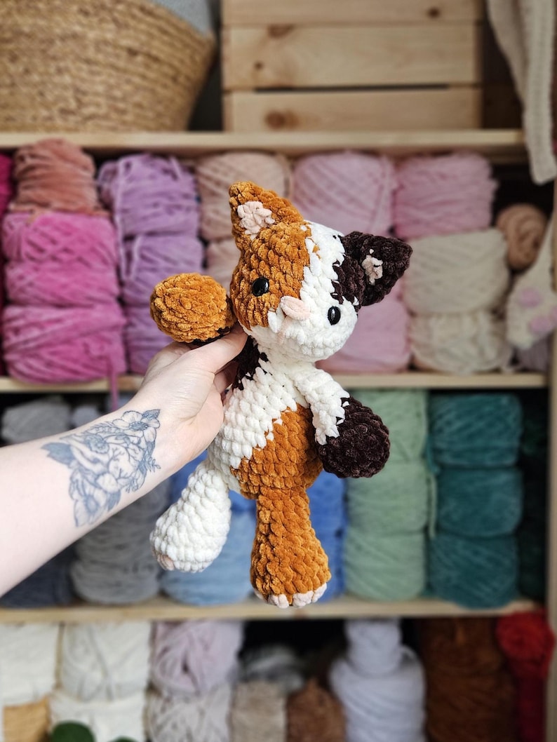 May include: A handmade crochet cat plushie in shades of brown, white, and dark brown. The plush toy has a soft, textured appearance and features a detailed design with small black eyes and a pink nose. The cat is held in front of a shelf filled with yarn.