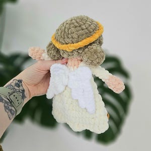 May include: A handmade crochet angel doll with a cream-colored dress and white wings. The doll has a brown head with a yellow halo. The arms and face are a light peach color. The doll is being held by a person.