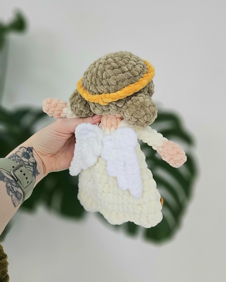 May include: A handmade crochet angel doll with a cream-colored dress and white wings. The doll has a brown head with a yellow halo. The arms and face are a light peach color. The doll is being held by a person.