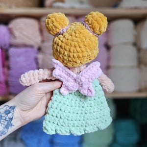 May include: A handmade crochet doll with a yellow head, pink bow, and a light green dress. The doll has two yellow pom-pom ears and is made of soft, textured yarn. The doll is a cute and cuddly toy.