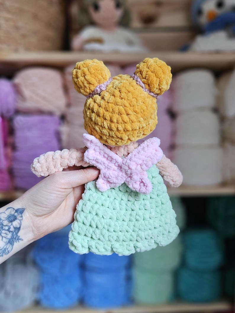 May include: A handmade crochet doll with a yellow head, pink bow, and a light green dress. The doll has two yellow pom-pom ears and is made of soft, textured yarn. The doll is a cute and cuddly toy.