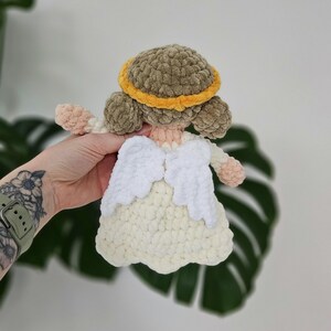 May include: A handmade crochet angel doll with a light brown halo, matching hair, and a cream-colored dress. The angel has white wings and peach-colored arms and hands. The doll is made of soft, textured yarn.