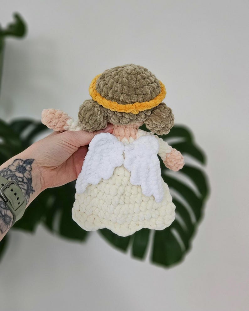May include: A handmade crochet angel doll with a light brown halo, matching hair, and a cream-colored dress. The angel has white wings and peach-colored arms and hands. The doll is made of soft, textured yarn.