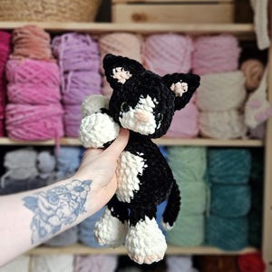 May include: A handmade black and white crochet cat doll with pink accents on the ears and nose. The plush toy is held in front of a shelf filled with colorful yarn skeins. The cat has green eyes and a small tail.