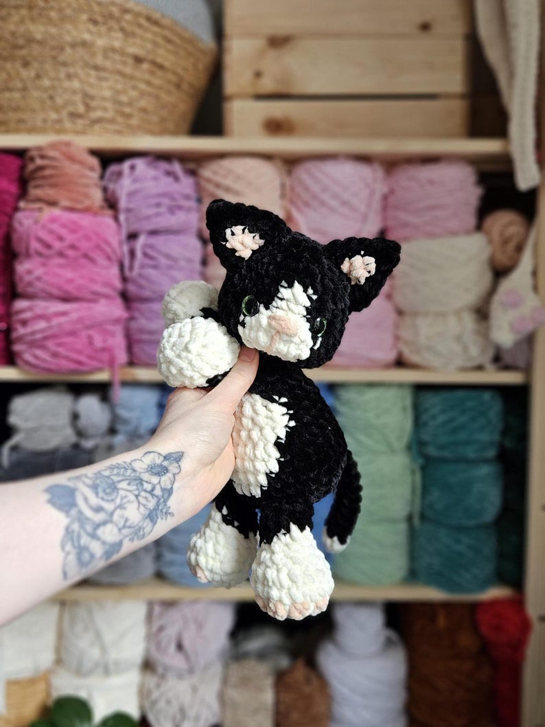 May include: A handmade black and white crochet cat doll with pink accents on the ears and nose. The plush toy is held in front of a shelf filled with colorful yarn skeins. The cat has green eyes and a small tail.