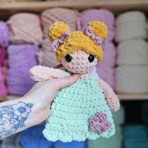 May include: A handmade crochet doll with a light green dress and a pink flower accent. The doll has yellow hair styled in two buns with pink bows. The doll's face is light pink with black eyes. The doll is being held in front of a blurred background of yarn.