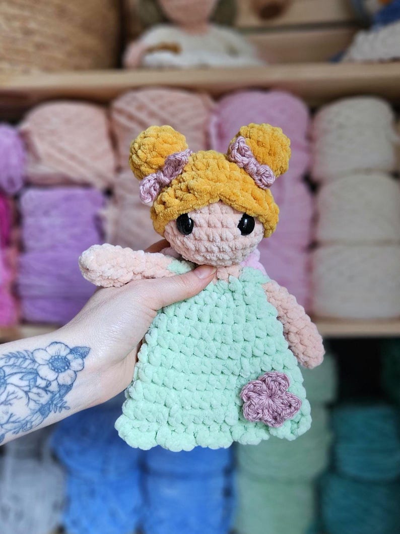 May include: A handmade crochet doll with a light green dress and a pink flower accent. The doll has yellow hair styled in two buns with pink bows. The doll's face is light pink with black eyes. The doll is being held in front of a blurred background of yarn.