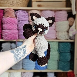 May include: A handmade crochet cat plushie in brown, white, and light blue. The cat has dark brown paws, ears, and facial markings, with light blue eyes and a white muzzle. The plushie is held in front of a shelf filled with yarn.